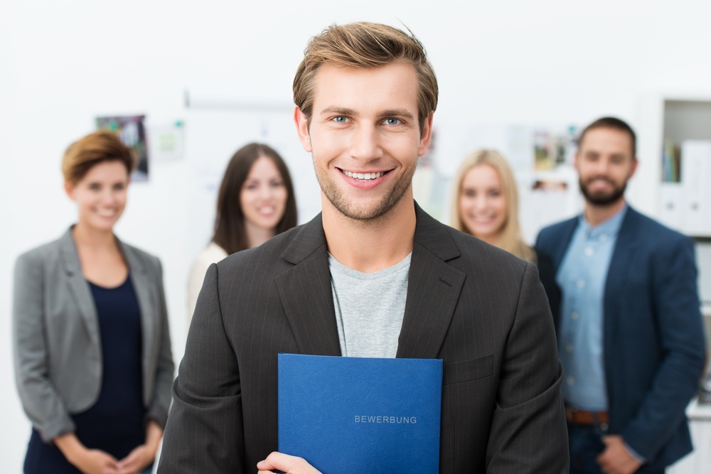 Successful smiling young male job applicant holding a blue file with his curriculum vitae posing in front of his new work colleagues or business team.jpeg